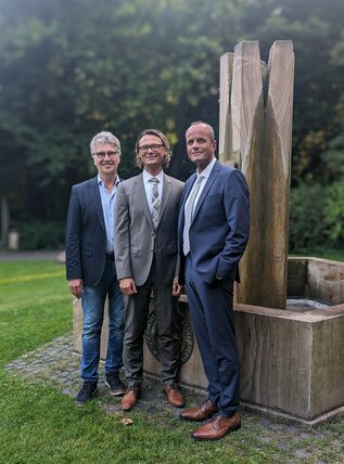 Drei Männer stehen vor einem Brunnen.